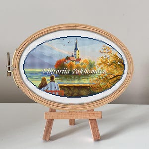 Könnte beinhalten: Ovale Kreuzstich-Stickerei einer Szene am Seeufer mit einer Kirche und einem Paar, eingefasst in einen Holzrahmen. Die Stickerei weist eine lebendige Farbpalette aus Blau-, Grün-, Orange- und Gelbtönen auf. Der Name des Künstlers, 'Viktoriia Pakhomova', ist unten aufgestickt.