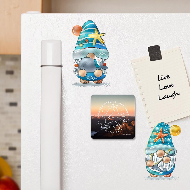 May include: A collection of decorative magnets on a white refrigerator. The magnets include two gnome designs with nautical themes, a square magnet with a mountain landscape, and a notepad with the words "Live Love Laugh".