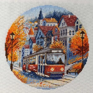 May include: A cross-stitch of a city scene with a red and white trolley car on tracks. The scene is framed by a circle of orange and yellow autumn foliage. Buildings with red and orange roofs are in the background, under a blue sky.