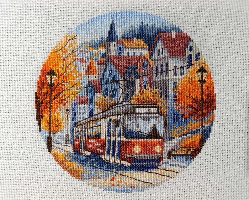 May include: A cross-stitch of a city scene with a red and white trolley car on tracks. The scene is framed by a circle of orange and yellow autumn foliage. Buildings with red and orange roofs are in the background, under a blue sky.