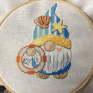May include: Cross-stitch embroidery of a gnome with a beach theme. The gnome wears a blue and yellow hat, holds a life preserver, and has a seashell and starfish on the hat. The embroidery is in a wooden hoop.