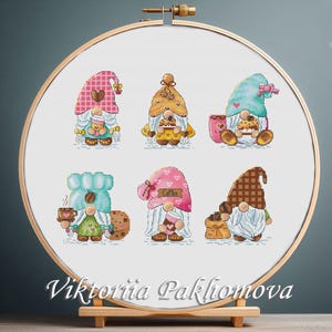 Puede incluir: Bordado de punto de cruz con seis gnomos con accesorios de café. Cada gnomo lleva un sombrero estampado y sostiene tazas de café y galletas. El bordado está en un aro de madera, con el nombre del artista en la parte inferior.