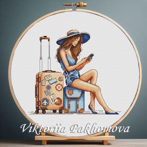 May include: Cross-stitch embroidery of a woman in a blue outfit, hat, and heels, sitting on a suitcase and using a phone. A second suitcase is next to her. The embroidery is in a wooden hoop. The text "Viktoriia Pakhomova" is at the bottom.