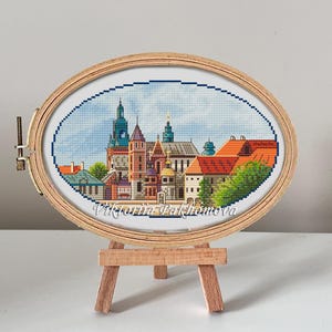 May include: A cross-stitch embroidery of a cityscape with a church and other buildings, framed in a wooden oval hoop and displayed on a wooden easel. The embroidery is signed "Viktoria Pakhomova".