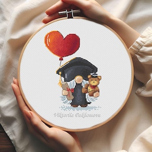 May include: A cross-stitch pattern featuring a gnome wearing a graduation cap and gown, holding a diploma and a teddy bear. A red heart-shaped balloon floats above the gnome. The pattern is framed in a wooden hoop and the text "Viktorija Pakhomova" is printed below the gnome.