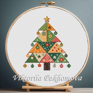 May include: A cross-stitch Christmas tree design in a wooden embroidery hoop. The tree is composed of colorful triangles with ornaments and a star. The design includes green, orange, red, and white colors. The artist's name, Viktoriia Pakhomova, is at the bottom.