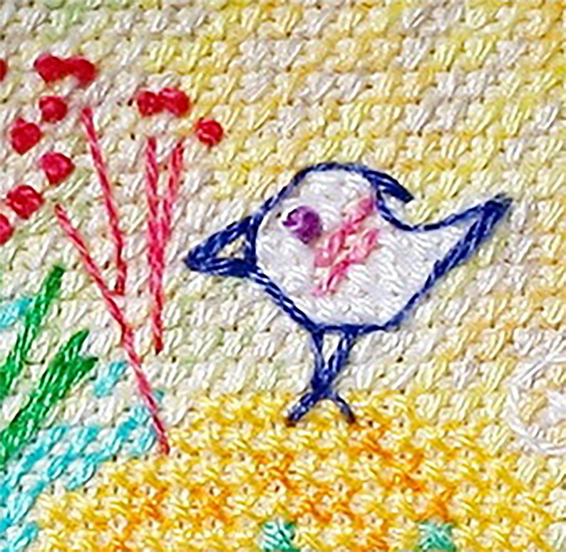 Op de afbeelding: Geborduurde vogel met blauwe vleugels en een wit lichaam, gepercht op een gele achtergrond met roze en rode bloemen.