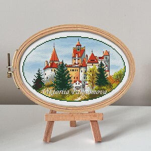 May include: A cross-stitch embroidery of a castle with red roofs and green trees, framed in a wooden oval hoop and displayed on a wooden easel. The embroidery is signed "Viktoria Pakhomova".