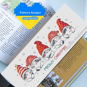Op de afbeelding: Een geborduurd boekenlegger met vier slapende katten met kerstmutsen. De tekst "MEOWY CHRISTMAS" is zichtbaar. De boekenlegger ligt op een open boek. De tekst "Pattern Keeper compatible" is ook zichtbaar.