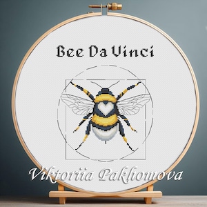 May include: Cross-stitch embroidery of a bee with yellow, black, and white details, framed in a wooden hoop. The text "Bee Da Vinci" is above the bee, and the artist's name, "Viktoriia Pakhomova," is below.