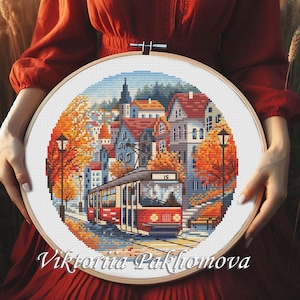 May include: A cross-stitch embroidery hoop featuring a detailed cityscape scene with a red tram, buildings, and autumn foliage. The artwork is set within a wooden hoop and held against a red background. The artist's name, Viktoriia Pakhomova, is at the bottom.