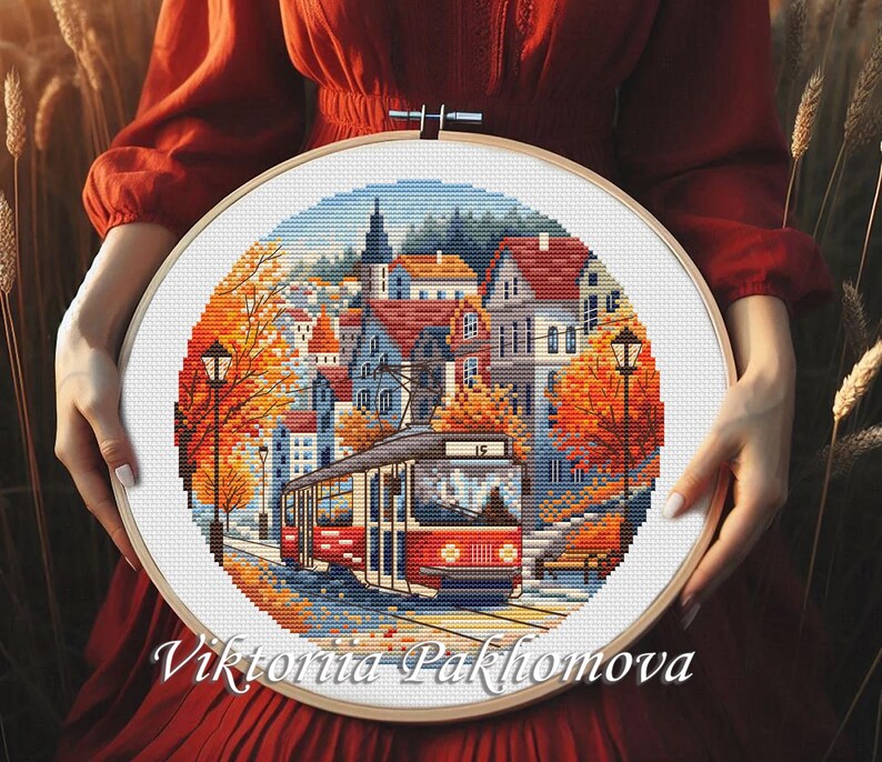 May include: A cross-stitch embroidery hoop featuring a detailed cityscape scene with a red tram, buildings, and autumn foliage. The artwork is set within a wooden hoop and held against a red background. The artist's name, Viktoriia Pakhomova, is at the bottom.