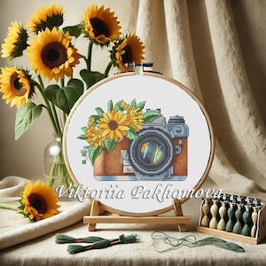 Könnte beinhalten: Kreuzstich-Stickerei einer Vintage-Kamera mit Sonnenblumen, präsentiert in einem Holzreifen. Die Kamera ist braun und blau, mit einem schwarzen Objektiv. Die Stickerei befindet sich auf weißem Hintergrund, mit dem Text "Viktoriia Pakhomova". Sonnenblumen sind in einer Vase.