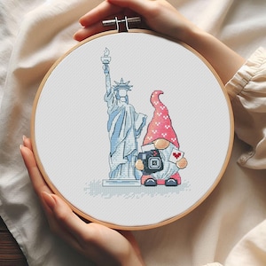 May include: Cross-stitch embroidery in a wooden hoop featuring the Statue of Liberty and a gnome with a camera and a heart-shaped card. The gnome wears a red hat with a white heart pattern and a red and white outfit. The Statue of Liberty is blue.