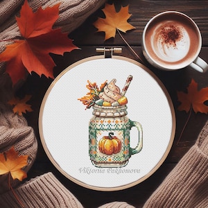 May include: Cross-stitch art in a wooden hoop depicts a fall-themed drink in a mason jar, topped with whipped cream and fall leaves. The jar features a pumpkin design. A cup of coffee and fall leaves surround the artwork.