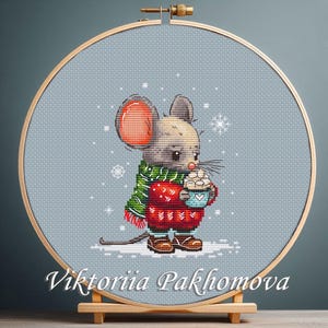 Może przedstawiać: Haft krzyżykowy przedstawiający mysz w czerwonym swetrze i zielonym szaliku, trzymającą niebieski kubek z piankami. Mysz ma duże uszy i jest umieszczona na jasnoniebieskim tle z płatkami śniegu. Tekst "Viktoriia Pakhomova" znajduje się na dole.