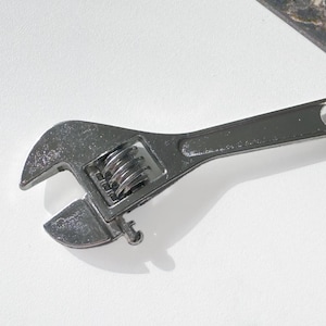 May include: A silver adjustable wrench keychain. The wrench is detailed with a movable jaw and a screw adjustment. It is attached to a chain and a split ring for keys. The background is white and a marbled surface.