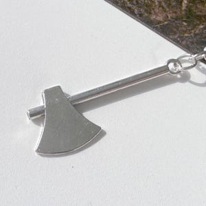 May include: A silver-colored axe keychain. The axe head and handle are made of metal and attached to a chain and a round key ring. The keychain is shown on a white and brown marble surface.