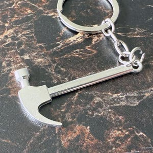 May include: A silver-colored hammer keychain with a ring and chain. The hammer has a curved claw and a flat head. The keychain is made of metal and is designed to be attached to keys or a bag.