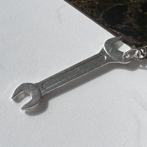 May include: A silver wrench keychain. The keychain features a small, detailed wrench charm attached to a chain and a round key ring. The wrench is a classic open-end design, and the metal has a shiny finish. The background is a mix of white and dark marble.
