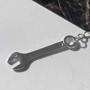 May include: A silver wrench keychain. The keychain features a miniature wrench with a loop for attaching keys. The wrench is made of shiny metal and has a detailed design. The keychain is on a white and brown surface.