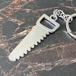 May include: A silver-colored saw-shaped keychain with a ring and chain. The saw blade has a series of triangular teeth. The keychain is made of metal and is designed to be attached to keys or a bag.