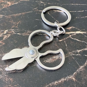 May include: Silver-colored keychain featuring a miniature scissors design. The scissors have a detailed, decorative shape and are attached to a split ring with a chain. The keychain is made of metal and is shown on a dark, marbled surface.
