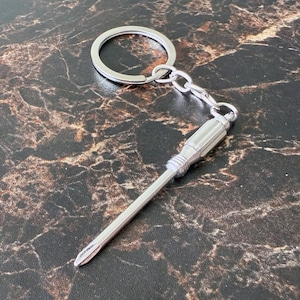 May include: A silver-colored screwdriver keychain with a Phillips head. The keychain includes a split ring and a short chain connecting to the screwdriver. The screwdriver has a textured handle and a pointed tip.