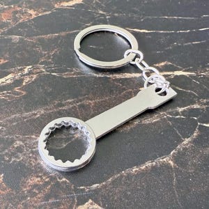 May include: A silver-colored keychain featuring a wrench-shaped charm. The charm has a circular end with a serrated interior and a flat, rectangular handle. The keychain includes a split ring and a short chain.