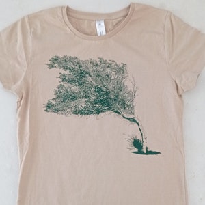 Könnte beinhalten: Ein beiges T-Shirt mit einer grünen Silhouette eines Baumes, der im Wind weht.