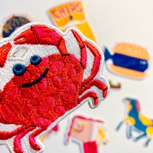 May include: A red embroidered crab patch with white stitching and blue eyes. The patch is on a white background with other embroidered patches in the background.