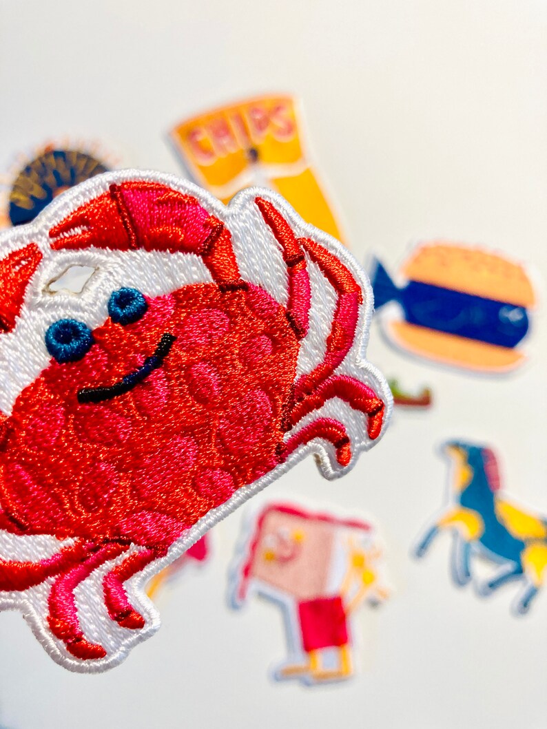 May include: A red embroidered crab patch with white stitching and blue eyes. The patch is on a white background with other embroidered patches in the background.