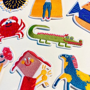 May include: A set of colourful embroidered iron-on patches featuring a variety of animals and objects, including a pufferfish, a bag of chips, a crab, a crocodile, a house, a horse, and a fish.
