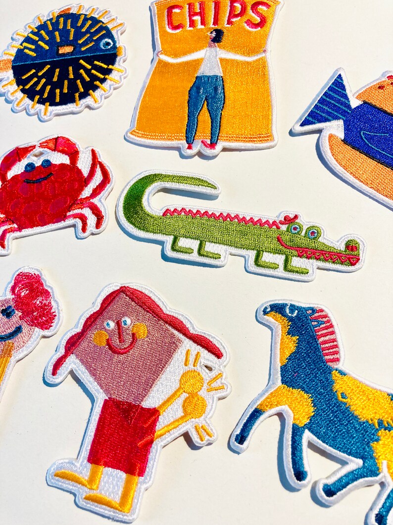 May include: A set of colourful embroidered iron-on patches featuring a variety of animals and objects, including a pufferfish, a bag of chips, a crab, a crocodile, a house, a horse, and a fish.