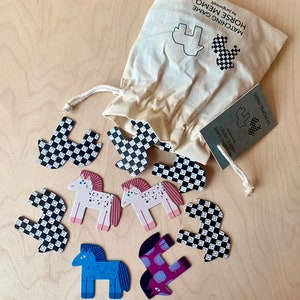 Könnte beinhalten: Ein Memory-Spiel mit 24 Pferdepaaren. Das Spiel beinhaltet eine Stofftasche mit einem schwarz-weiß karierten Pferde-Design. Die Spielsteine sind aus Holz und zeigen bunte Pferde mit verschiedenen Mustern.