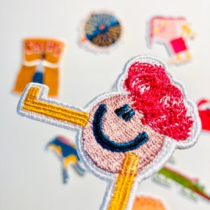 May include: An embroidered patch featuring a smiling face with red hair and a yellow stick figure body. The patch is surrounded by other colourful patches.