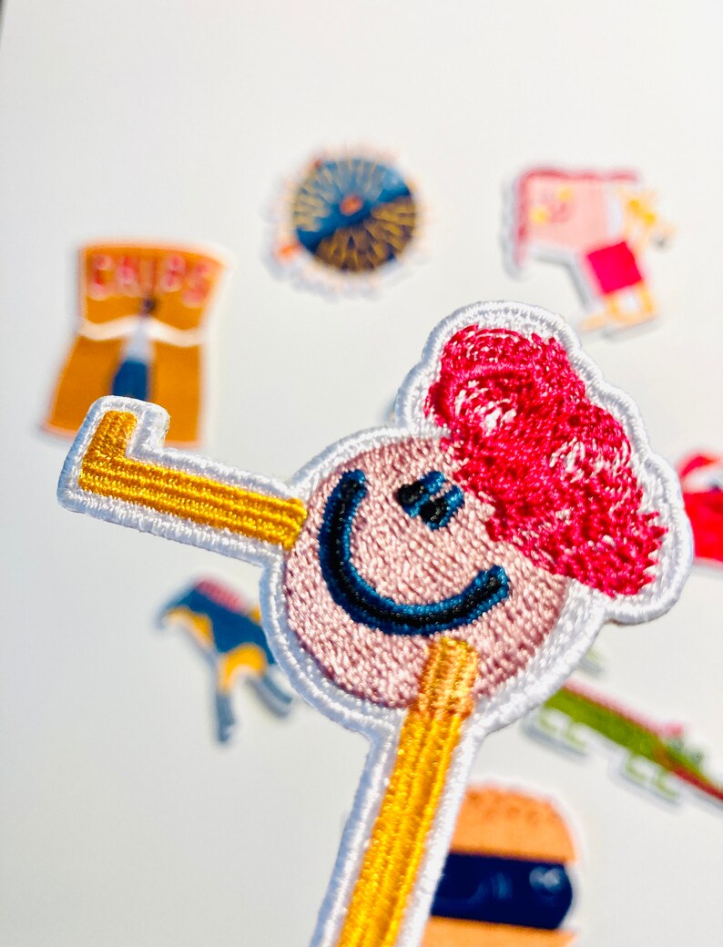 May include: An embroidered patch featuring a smiling face with red hair and a yellow stick figure body. The patch is surrounded by other colourful patches.