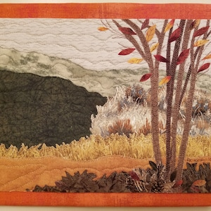 May include: A quilted wall art piece depicting a fall landscape with a brown tree with red and yellow leaves in the foreground, a field of tall grass, and a mountain range in the background.