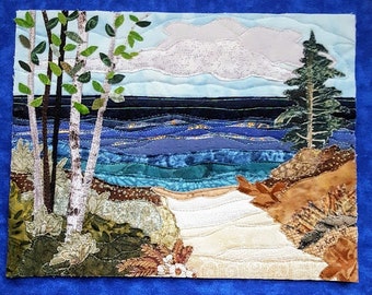 Puede incluir: Una obra de arte textil que representa un paisaje costero. La obra presenta un océano azul, un cielo nublado y una costa con árboles. La pieza está enmarcada con un borde azul y muestra una variedad de texturas y colores.