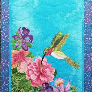 May include: A quilted wall hanging featuring a hummingbird in flight with a pink, purple, and green floral design on a turquoise background.