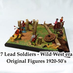 May include: A Wild West diorama with seven lead soldiers from the 1920s-50s. The scene includes miniature figures of cowboys and Native Americans, set amongst small trees, logs, and a wooden wagon wheel. The base is a dark, rectangular shape.