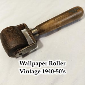 May include: Vintage wallpaper roller with a wooden handle and roller. The roller is dark brown, and the handle is a lighter wood tone. The metal frame is silver. The text "Wallpaper Roller Vintage 1940-50's" is below the roller.