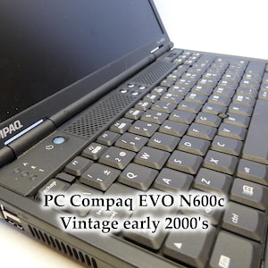 May include: A black Compaq EVO N600c laptop from the early 2000s. The laptop is open, displaying the keyboard and screen. The keyboard has black keys with white lettering. The laptop has a blue touchpad button and the word "COMPAQ" is visible.