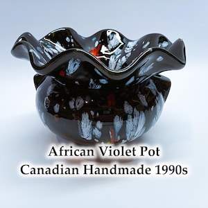 Op de afbeelding: Handgemaakte Afrikaanse viooltjespot uit de jaren 1990. De donkerbruine keramische pot heeft een glanzende afwerking en een gegolfde rand. Witte en blauwe verfspatten versieren de buitenkant, met rode accenten.