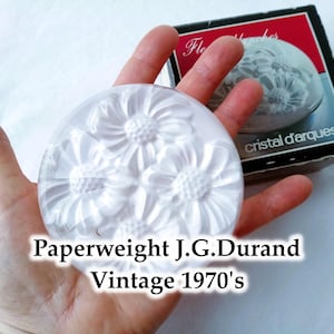 May include: A vintage 1970s J.G. Durand paperweight. The clear glass paperweight has a raised floral design. The round paperweight is next to its original box. The box has a red and black design with the words "cristal d'arques".