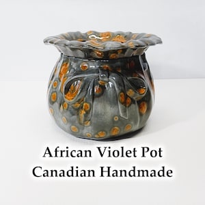 May include: Handmade African Violet Pot. The ceramic pot is grey with orange speckled accents and a ruffled rim. The pot has a bow design on the front. The text "African Violet Pot" and "Canadian Handmade" are at the bottom.