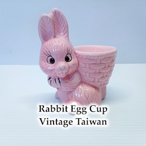 Peut inclure: Un coquetier en céramique rose en forme de lapin avec un porte-œuf en forme de panier. Le lapin a des yeux noirs, des cils et un nœud papillon. Le texte "Rabbit Egg Cup Vintage Taiwan" est en dessous.