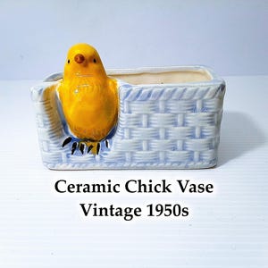 May include: A vintage ceramic vase from the 1950s, featuring a bright yellow chick inside a light blue basket-weave container. The rectangular vase is suitable for small plants or decorative purposes.