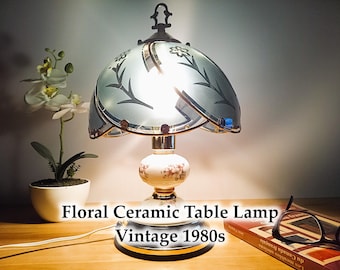May include: Vintage floral ceramic table lamp from the 1980s. The lamp features a floral patterned base and a light blue glass shade with floral and leaf designs. The lamp is turned on, illuminating the room.