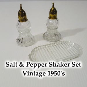Peut inclure: Ensemble de salière et poivrière vintage des années 1950. L'ensemble comprend deux salières en verre transparent avec des couvercles dorés et un plateau en verre transparent assorti. Le texte "Salt & Pepper Shaker Set Vintage 1950's" est visible.